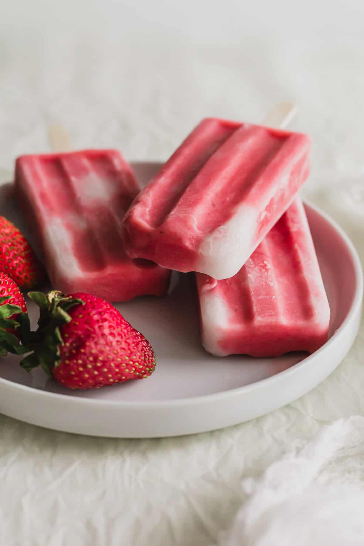 3 Strawberry sago popsicles piled on a white plate, with strawberries on the side.